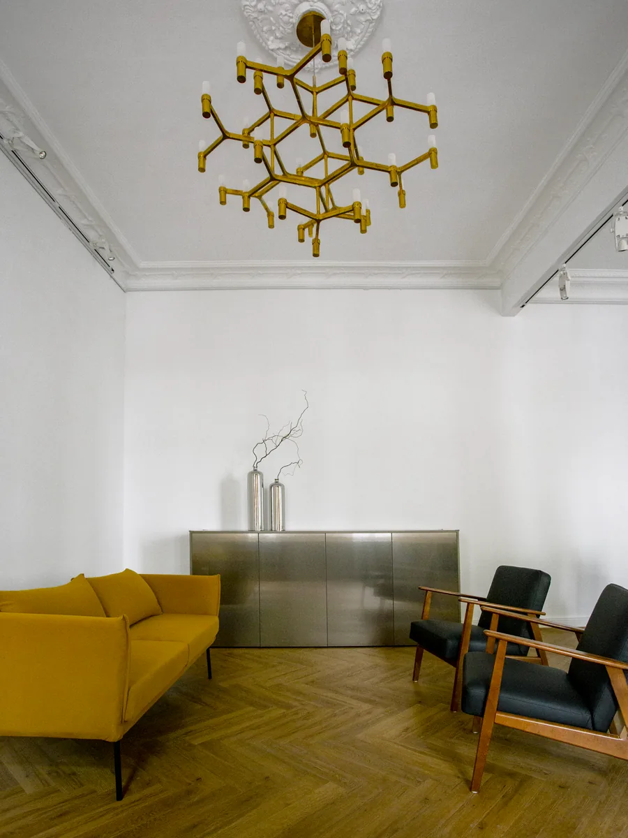 Ein Raum mit einem Messing-Kronleuchter an der Decke, einem gelben Sofa, einem Metallschrank und zwei Sesseln auf Fischgrätparkett