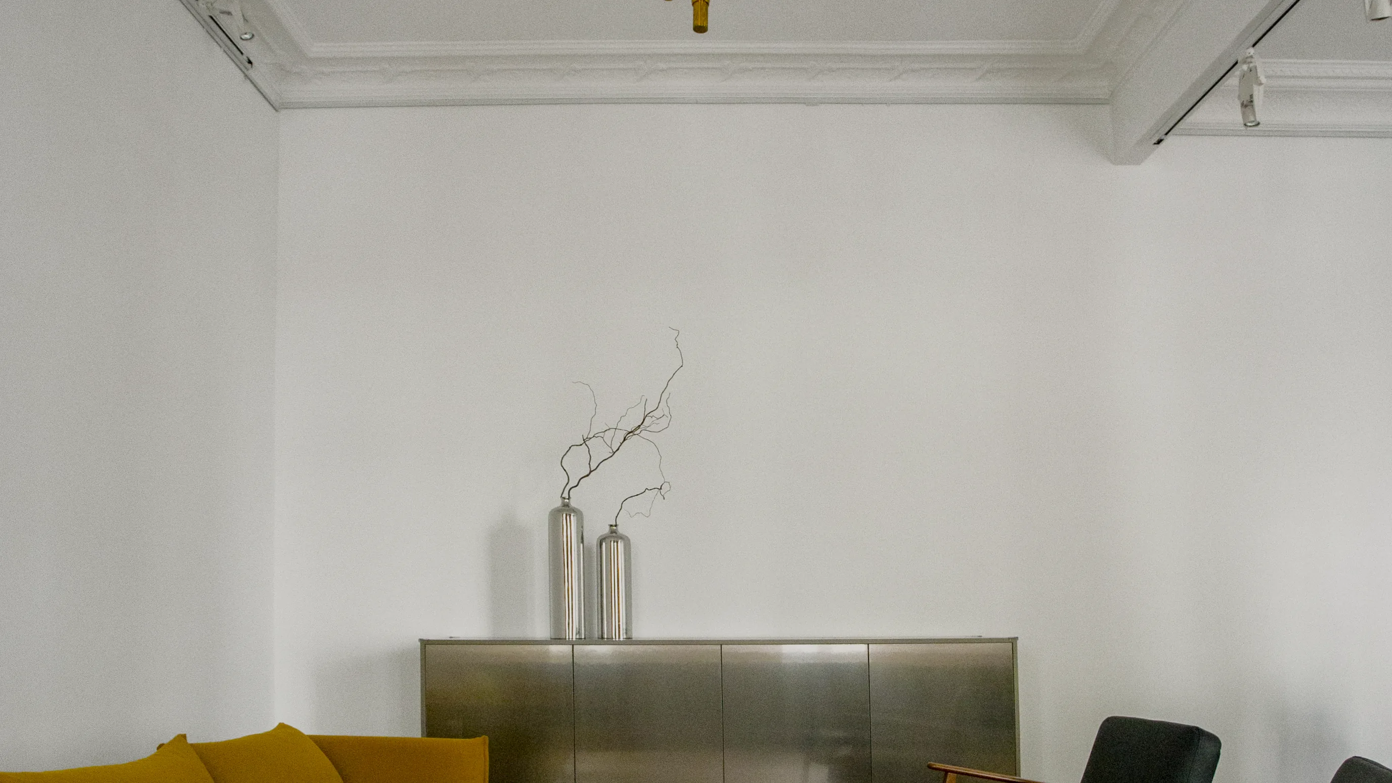 Ein Raum mit einem Messing-Kronleuchter an der Decke, einem gelben Sofa, einem Metallschrank und zwei Sesseln auf Fischgrätparkett