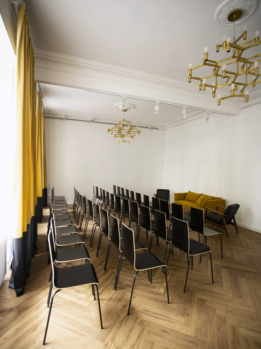 Stuhlreihen aus schwarzen Stühlen im Saal aus der Perspektive des Fensters, zwei Messing-Kronleuchter an der Decke, ein gelbes Sofa in der Ecke