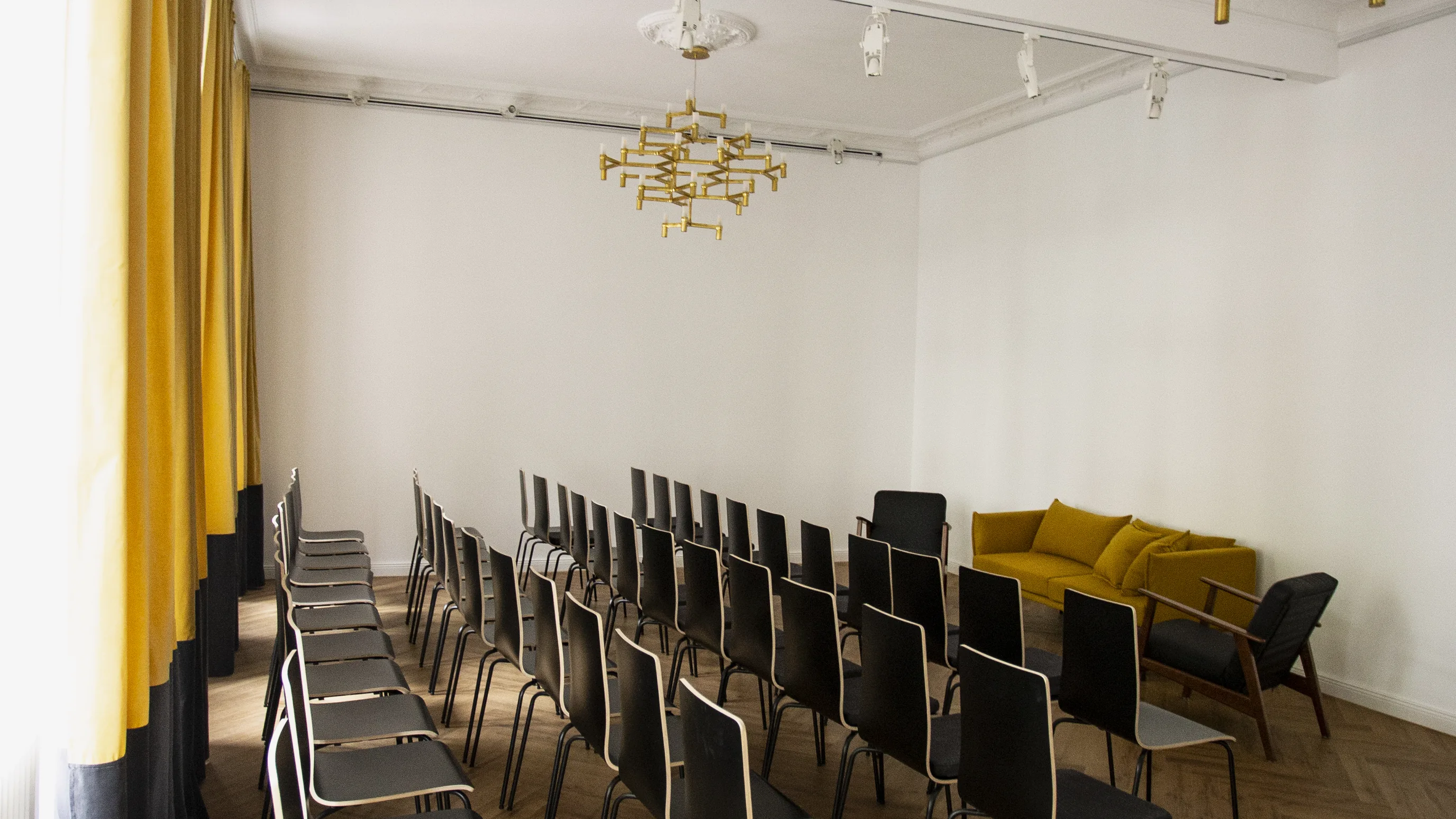 Stuhlreihen aus schwarzen Stühlen im Saal aus der Perspektive des Fensters, zwei Messing-Kronleuchter an der Decke, ein gelbes Sofa in der Ecke