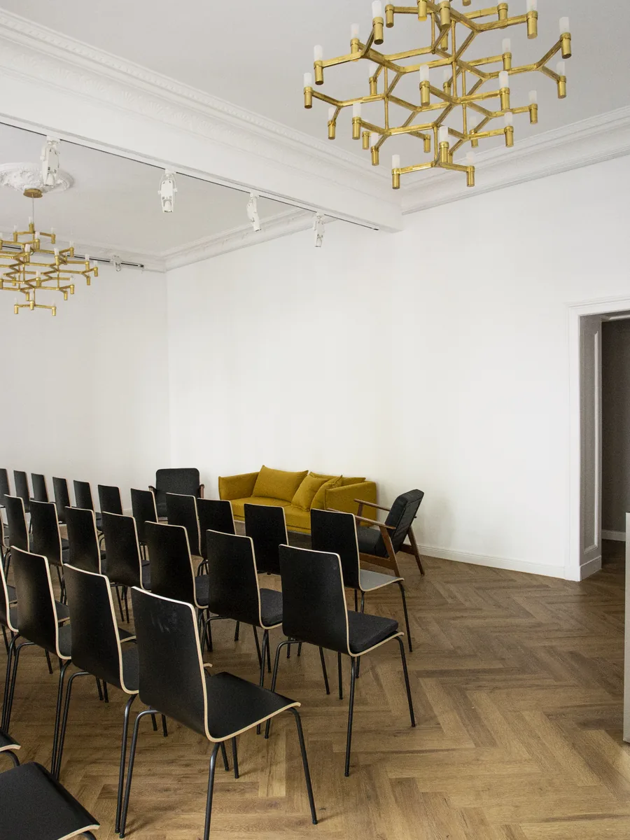 Stuhlreihen aus schwarzen Stühlen im Saal, ein gelbes Sofa im Hintergrund, rechts eine Metalltheke mit Laptop und Vasen