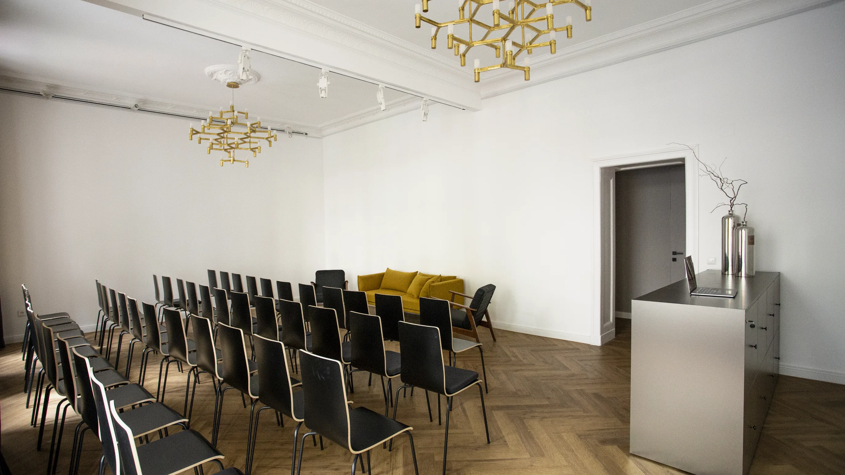 Stuhlreihen aus schwarzen Stühlen im Saal, ein gelbes Sofa im Hintergrund, rechts eine Metalltheke mit Laptop und Vasen
