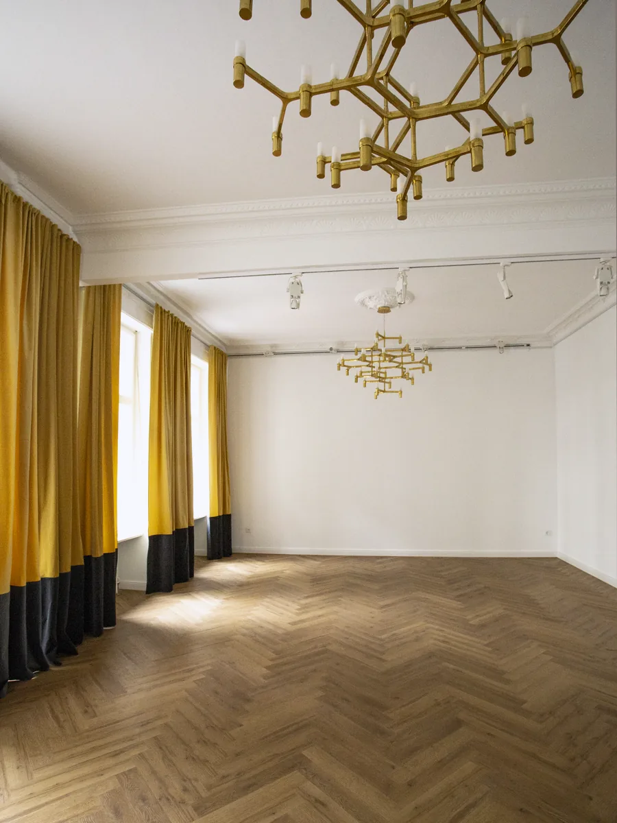 Ein leerer Saal ohne Möbel, Fischgrätboden, Vorhänge oben gelb und unten schwarz, zwei Messing-Kronleuchter an der Decke