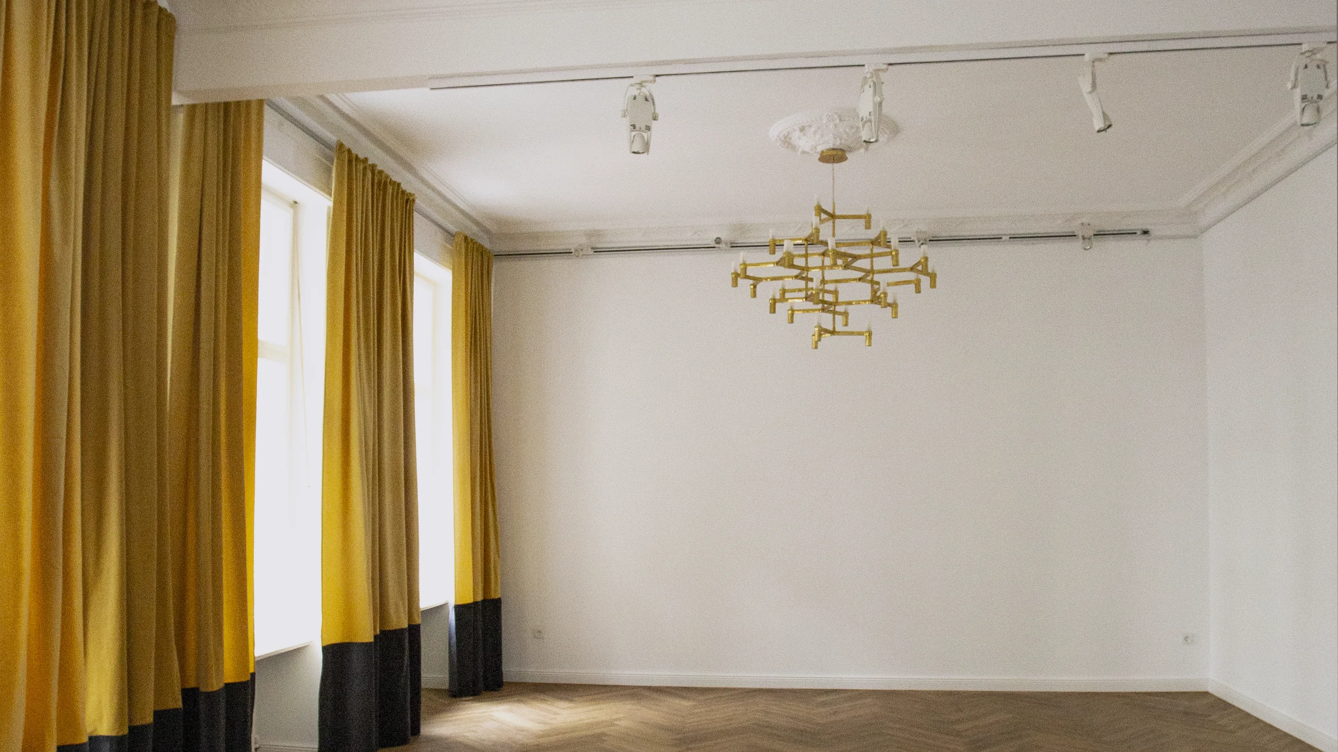 Ein leerer Saal ohne Möbel, Fischgrätboden, Vorhänge oben gelb und unten schwarz, zwei Messing-Kronleuchter an der Decke