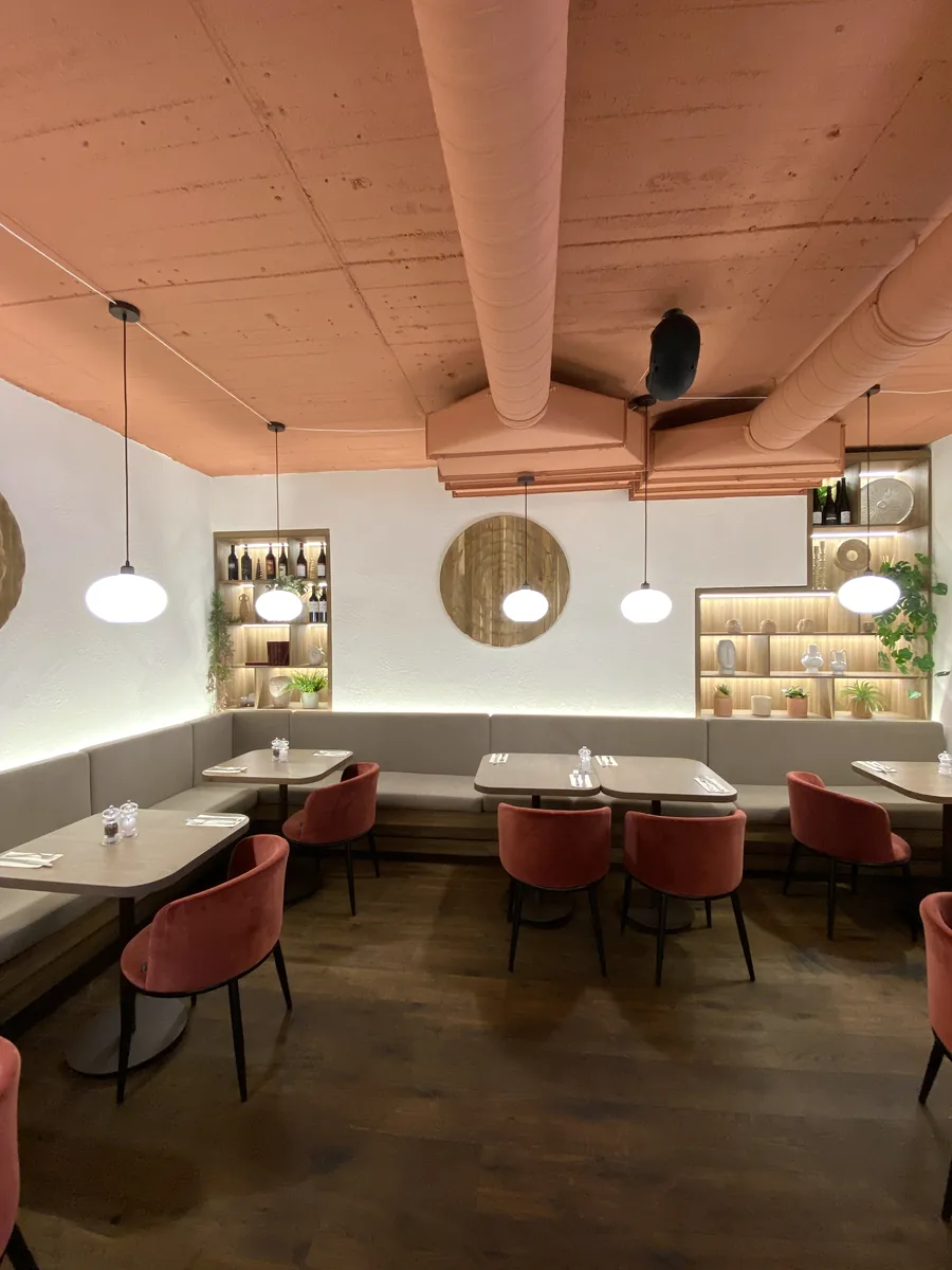 Café-Saal mit einer Tischreihe und einer Sitzbank entlang der Wand, in den Nischen über der Bank stehen Flaschen und Geschirr auf Regalböden, kugelförmige Hängeleuchten