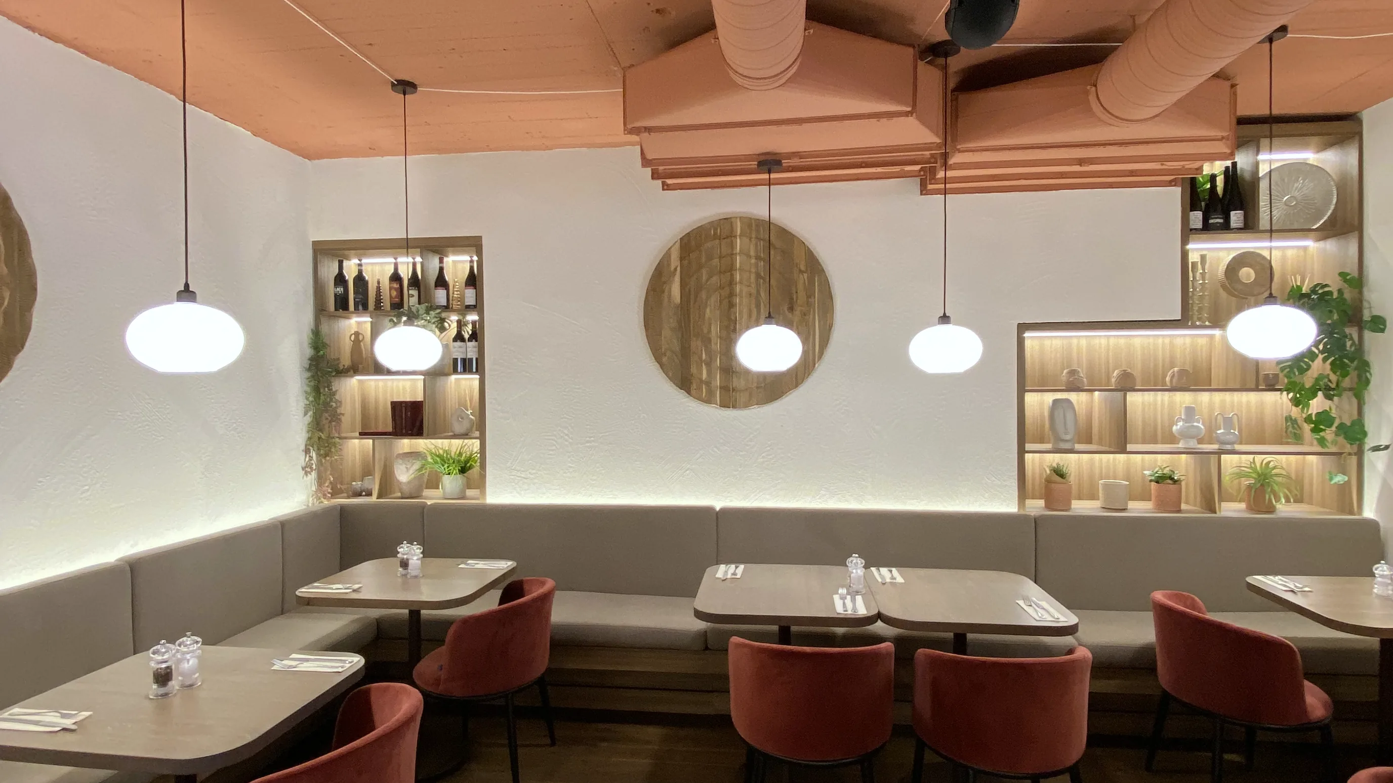Café-Saal mit einer Tischreihe und einer Sitzbank entlang der Wand, in den Nischen über der Bank stehen Flaschen und Geschirr auf Regalböden, kugelförmige Hängeleuchten