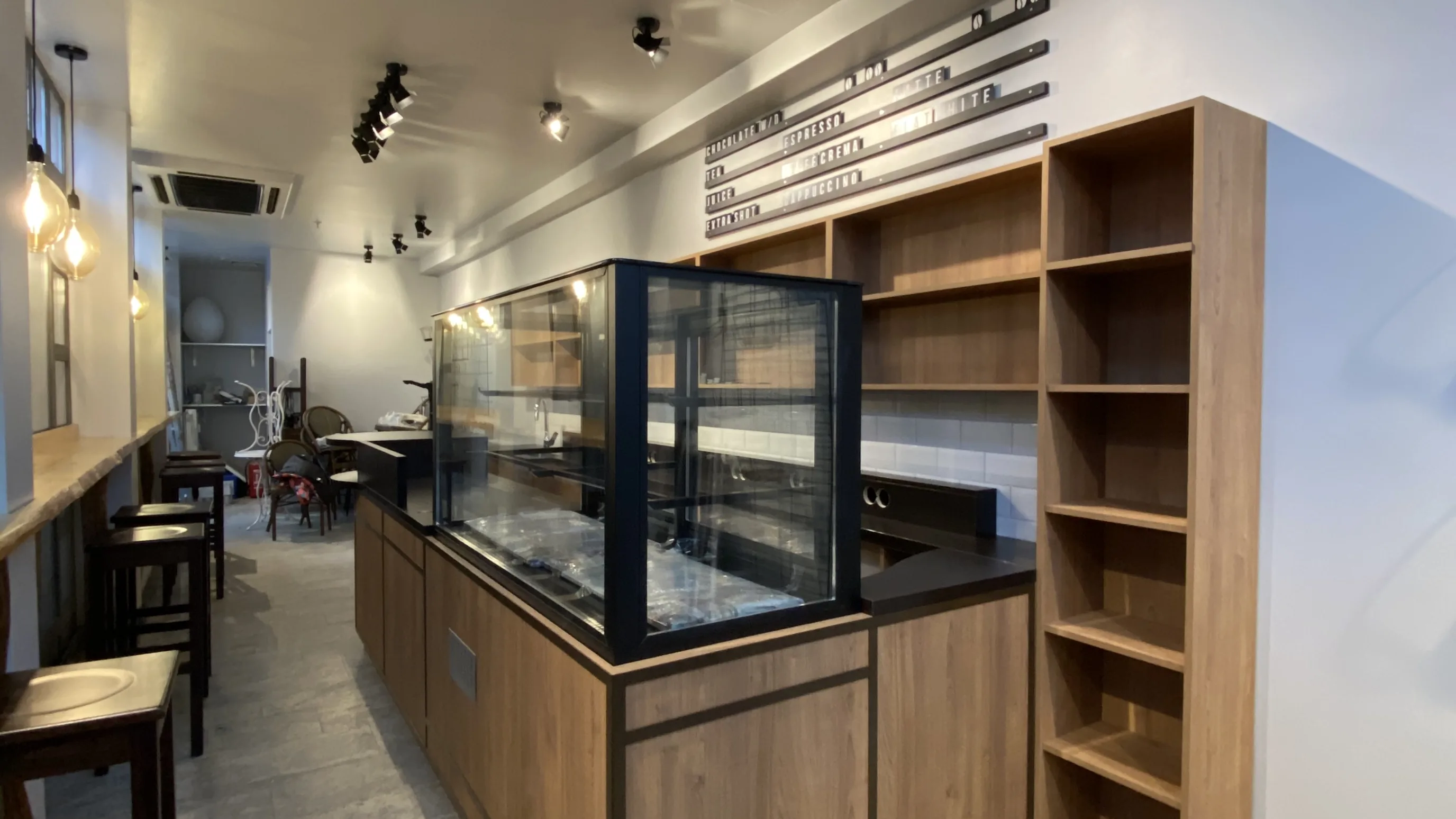 Langer schmaler Caféraum mit einer Holztheke und einer Glasvitrine in der Mitte, daneben offene Regale, im Hintergrund Tische und Barhocker
