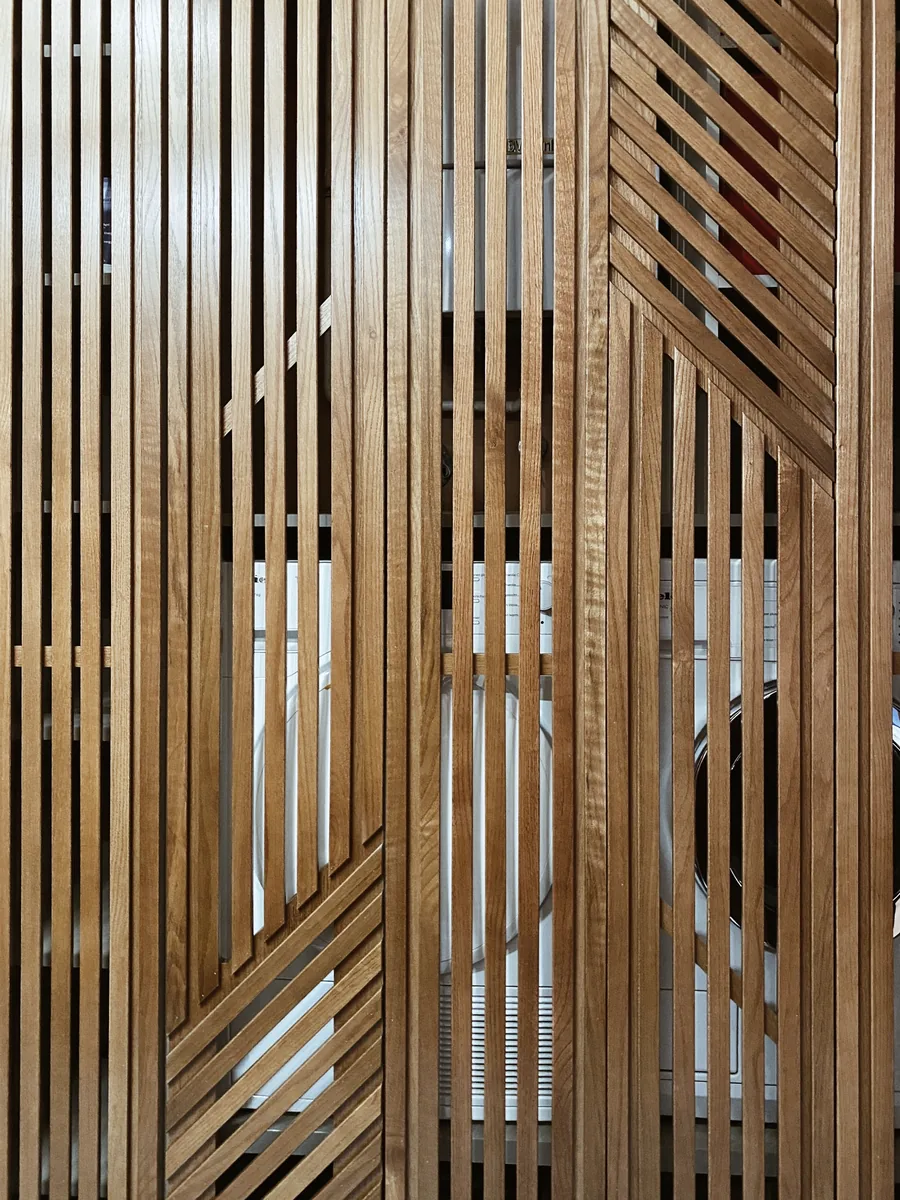 Nahaufnahme einer Front aus vertikalen Eichenlamellen in geometrischem Muster, hinter dem Glas sind Umrisse von Geräten zu erkennen