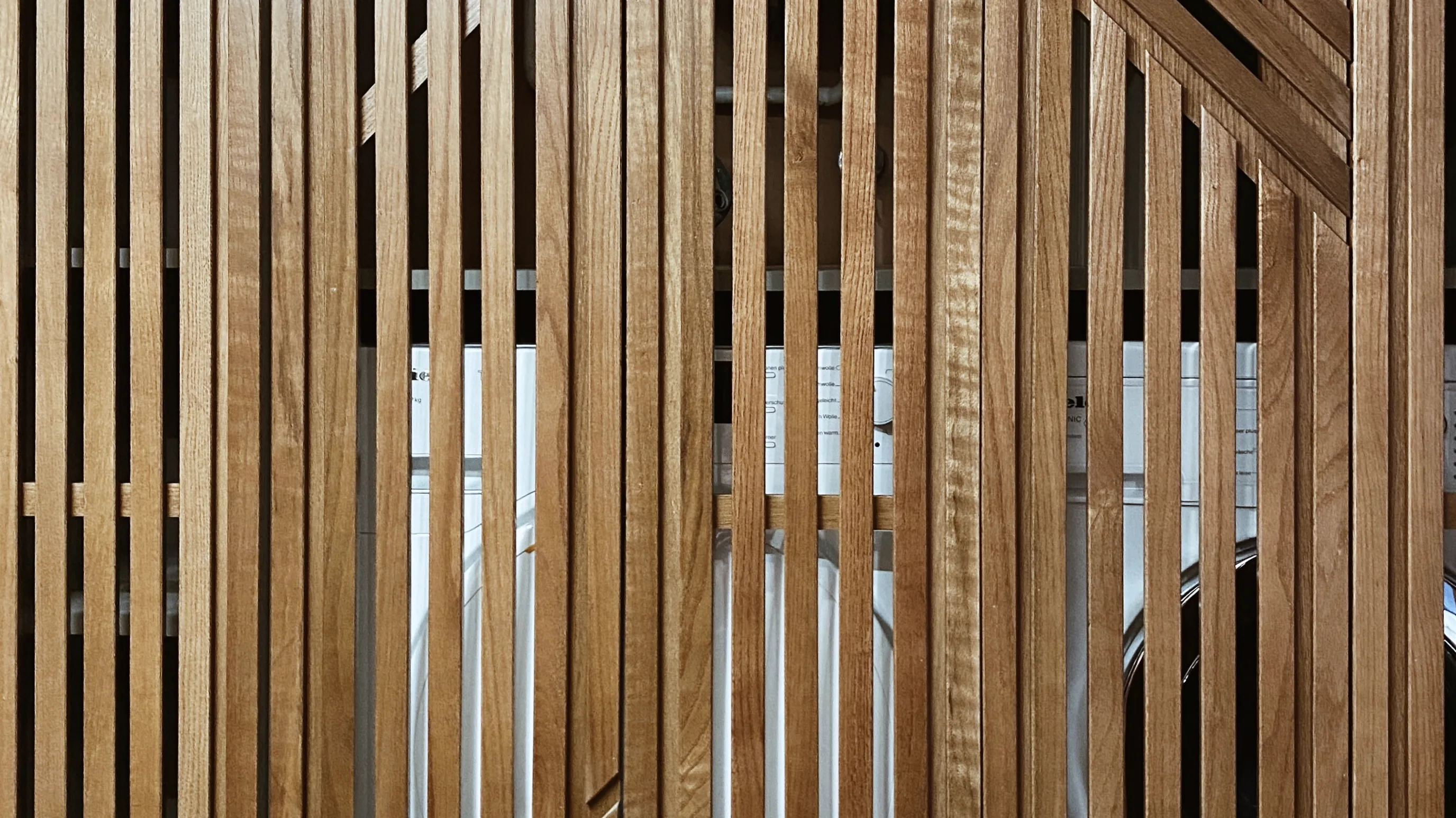 Nahaufnahme einer Front aus vertikalen Eichenlamellen in geometrischem Muster, hinter dem Glas sind Umrisse von Geräten zu erkennen
